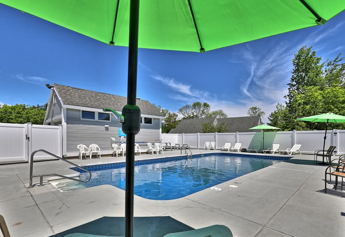 This image features a sunlit swimming pool surrounded by green umbrellas and lounge chairs within a fenced yard, all set against a relaxed outdoor backdrop. The inviting pool area hints at resort-style amenities and is ideal for summer enjoyment.