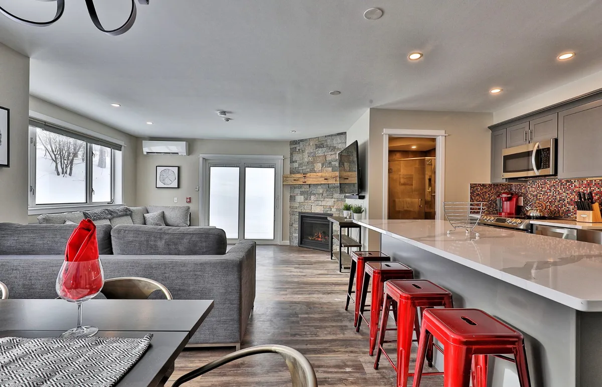 A bright, open-plan living area featuring a modern gray sectional sofa, a set of eye-catching red bar stools at the kitchen island, and a handsome stone fireplace. The space is designed for both comfort and entertaining, with sightlines that encourage group interaction and plenty of room for gathering.