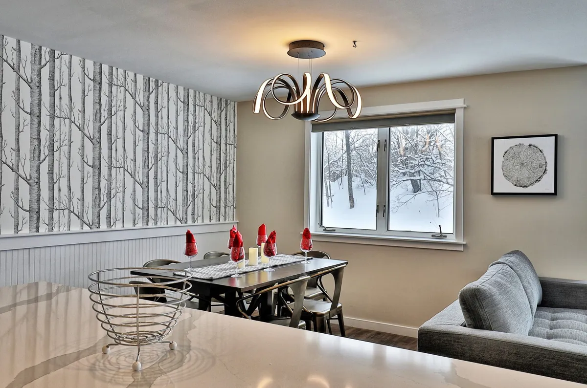 The image captures a dining area with a table set near a large window that frames a picturesque snowy landscape. The room is accented by tree-patterned wallpaper and a modern chandelier, creating an inviting space for meals or gatherings while watching snowflakes fall outside.