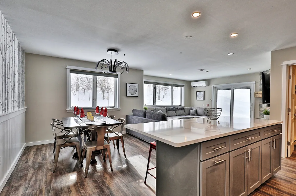 A bright, open dining area flows seamlessly from the living space, featuring a dining table with red accents and a plush gray sofa nearby. The large windows frame a snowy landscape, infusing the space with light and connecting the indoors with the outdoors. The room exudes warmth and is ideal for memorable meals or lively game nights.