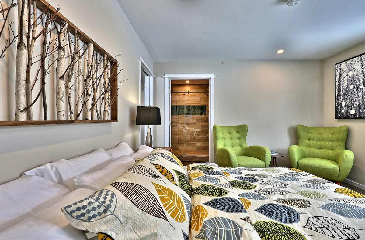 A tranquil bedroom with a king-sized bed adorned in patterned bedding, complemented by green accent chairs and wooden wall art featuring branches. Soft natural light fills the space, and nightstands with lamps provide a cozy touch. The room feels calm and inviting, perfect for relaxation and restful sleep.