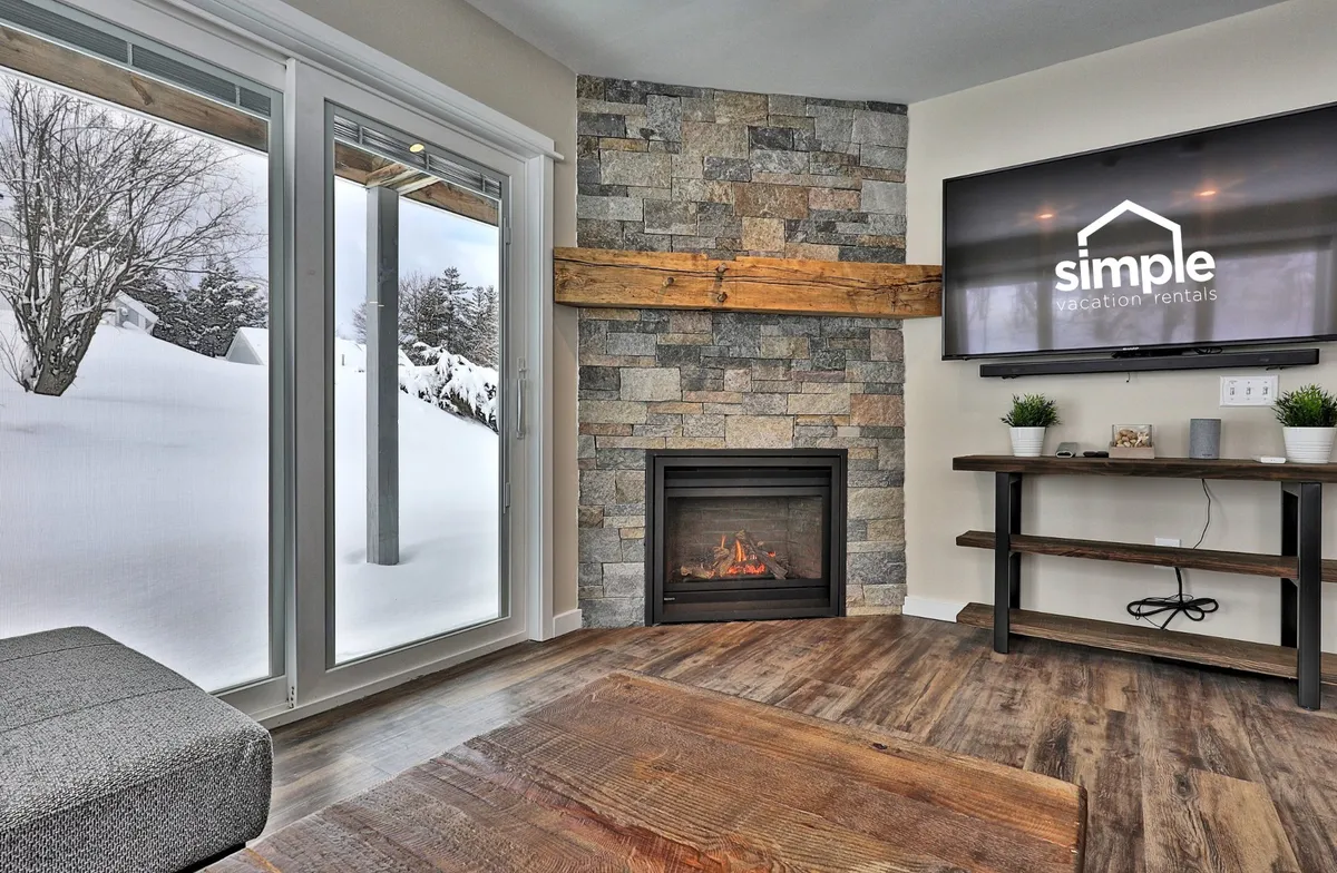 This image captures a warmly lit living room anchored by a striking fireplace. Large windows invite ample natural light and display tranquil snowy mountain views. There's a comfortable sofa arrangement, a TV for entertainment, and wood beams adding a cozy, rustic charm. The setting is perfect for relaxing by the fire, gathering for conversation, or enjoying the scenery.