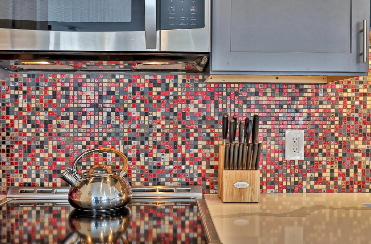 A stylish kitchen with a colorful mosaic backsplash, stainless steel kettle, and a block of knives on the counter. The lively setup hints at joyful meal preparation, cooking classes, or festive family dinners.
