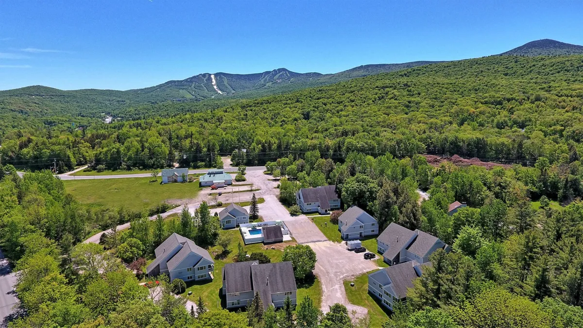 The image showcases a wide-open green field bordered by dense trees, with clusters of houses nestled at the base of majestic mountains. The peaceful expanse and inviting nature trails evoke a sense of escape and proximity to outdoor adventures.