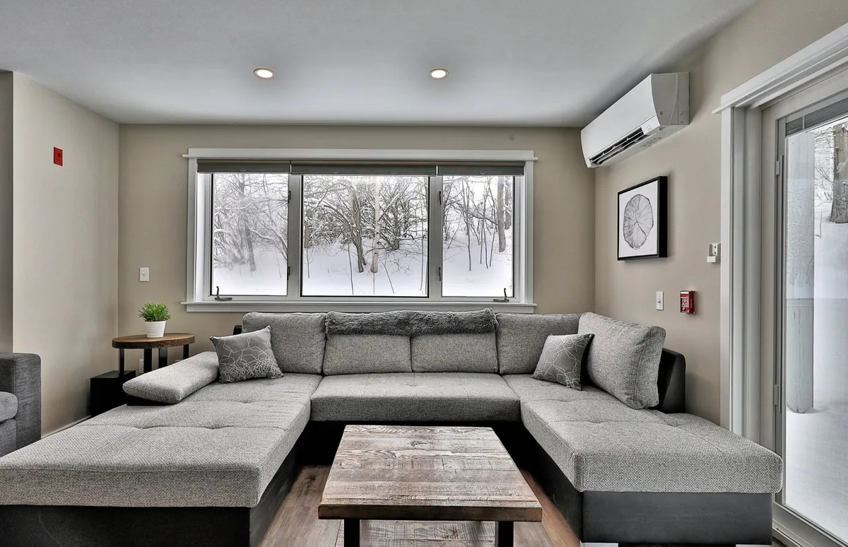 This interior image features a large sectional sofa positioned by windows that offer clear views of a snowy landscape. The cozy arrangement is perfect for relaxing, socializing, and watching snow fall from the comfort of the home. The warm, inviting atmosphere suggests a welcoming retreat after outdoor winter adventures.