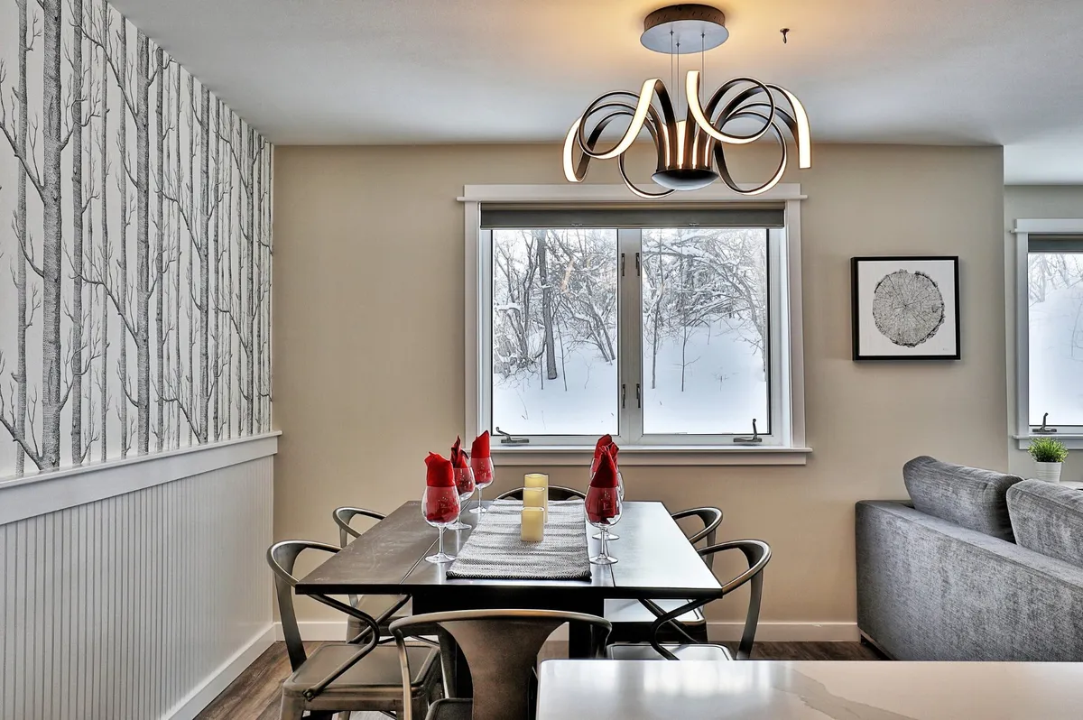 A cozy dining space with a wooden table under a sparkling chandelier, set against a wall decorated with a tree pattern and windows framing a snowy landscape. The setup invites shared meals and intimate celebrations, capturing the feeling of special occasions.
