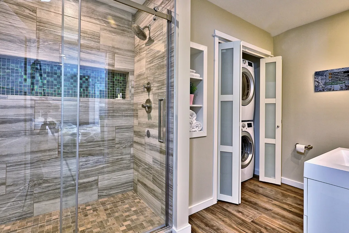 An efficient, modern laundry area with sleek washer/dryer units, glass shower enclosure, and textured wall tiles. The space is bright and practical, emphasizing convenience and ease for guests.