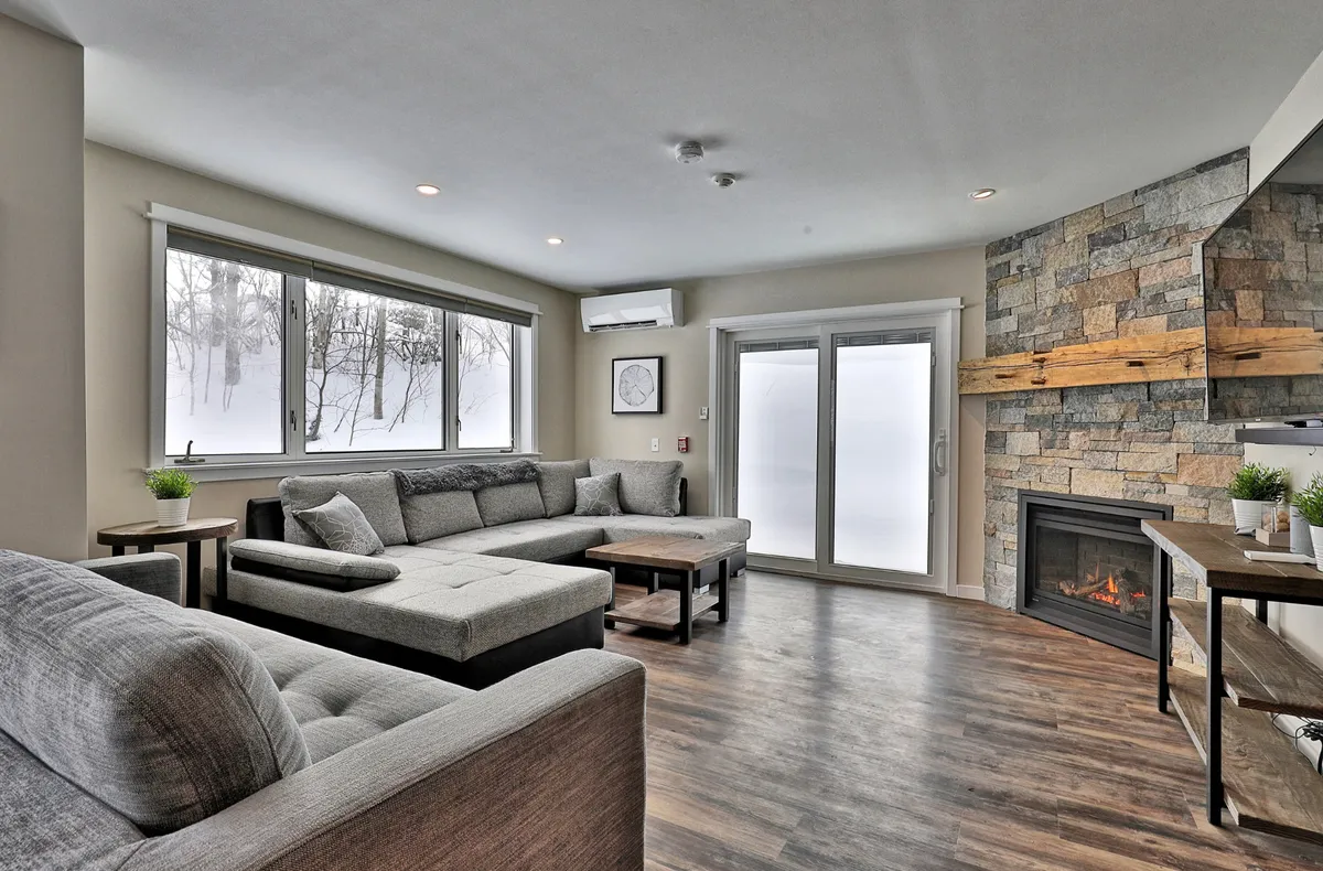 A welcoming living space with a fireplace as its focal point, surrounded by a comfortable sofa arrangement and large windows. The room is designed for relaxing, family conversations, and reading by the fire, especially as snow falls outside.