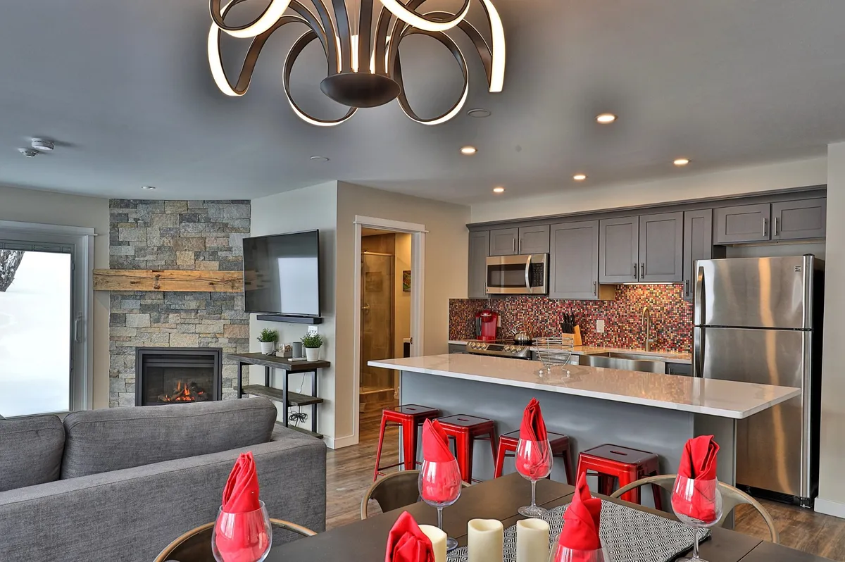 A stylish, renovated condo interior featuring a modern chandelier illuminating a spacious dining area set for a festive meal, a cozy stone fireplace inviting relaxation, and a vibrant backsplash highlighting the well-equipped kitchen. The scene exudes warmth and comfort—perfect for family gatherings or après-ski evenings with friends.