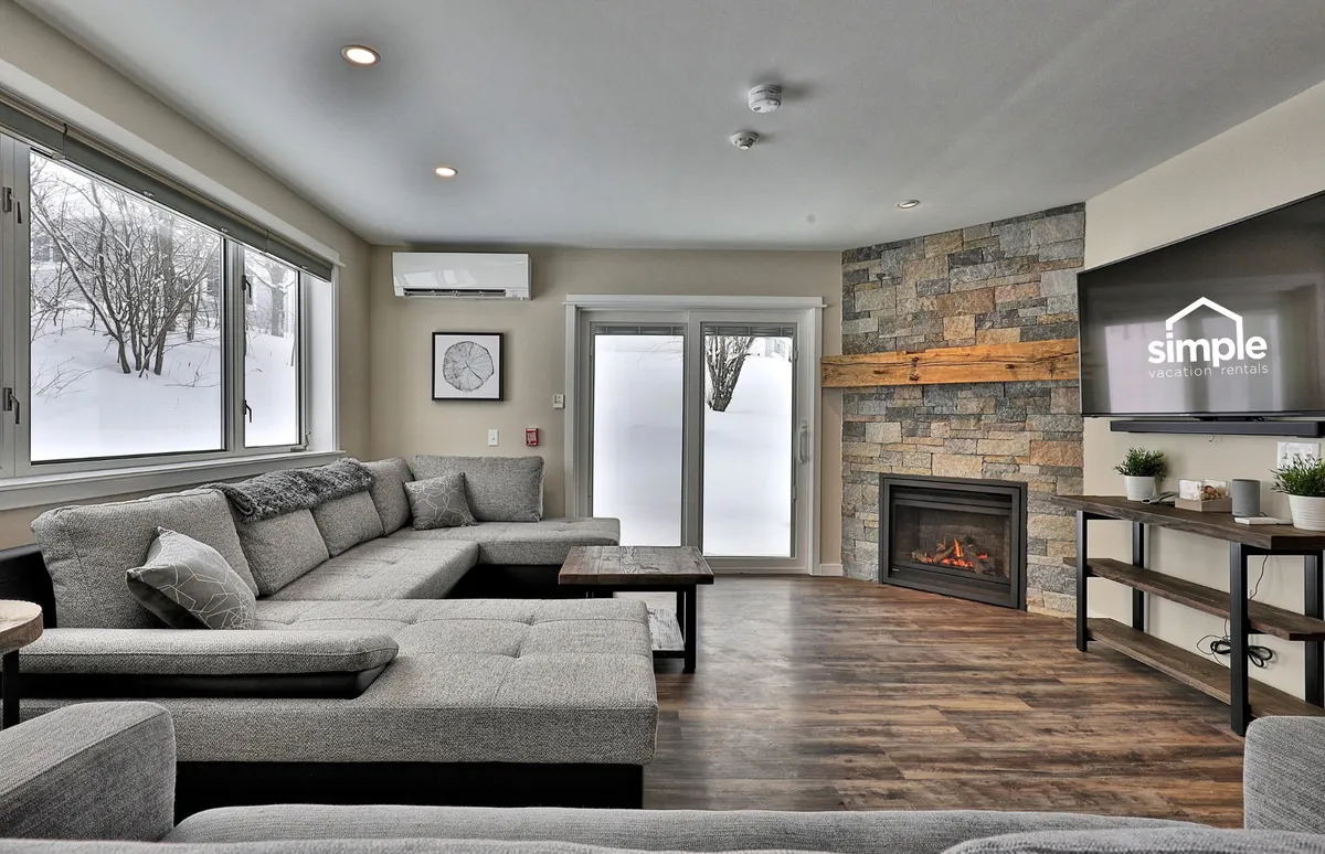 A spacious living room with a large sectional sofa faces a stone fireplace and television, all set against a backdrop of panoramic windows revealing the snowy outdoors. The space is designed for both entertaining and relaxing, offering comfortable seating for groups and an atmosphere perfect for movie nights or unwinding after outdoor adventures.